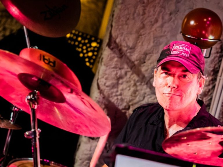 Drummer in zwart shirt en pet, spelend op een podium met kleurrijke verlichting.
