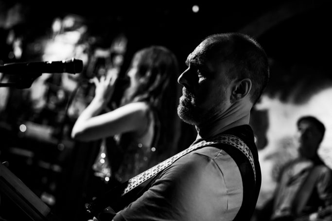 Zwart-wit afbeelding van een muzikant met een gitaar, terwijl een zangeres op de achtergrond zingt.