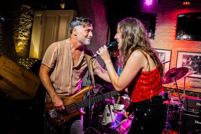 Zanger en zangeres optreden op een podium met instrumenten en gekleurde verlichting.