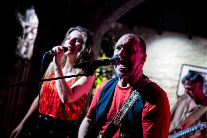 Een man en vrouw zingen samen op een podium, met een muzikant op de achtergrond.
