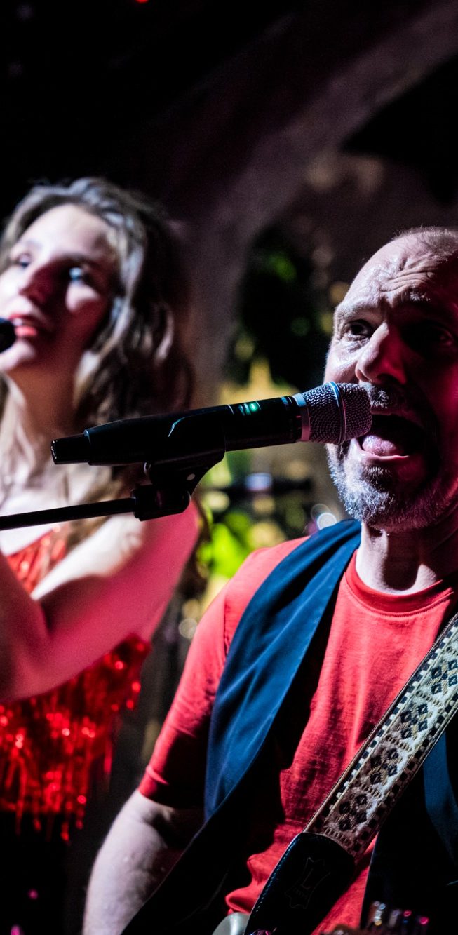 tribute band vrouwen, female rock tribute, live band Nederland, avondvullend, feestband Een man en vrouw optreden met microfoons in een levendige muzieksetting.