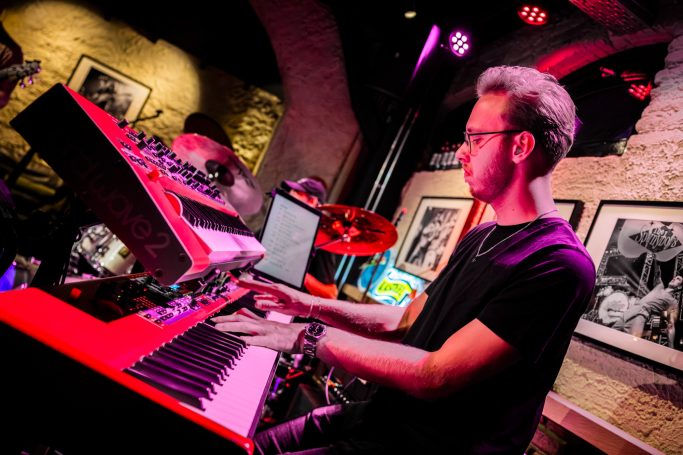 Man playing keyboards on stage with drums and colorful lighting in the background.