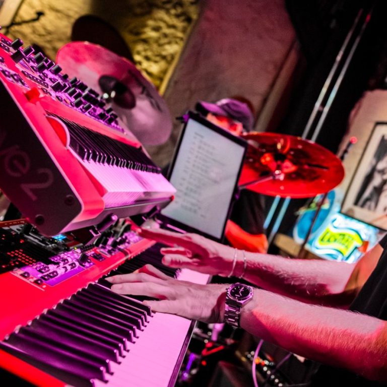 Musician playing a synthesizer and keyboard on stage, with drums in the background.