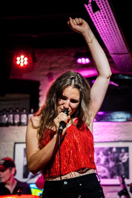 Zangeres met een mic, steekt haar arm op in een live muzieksetting.