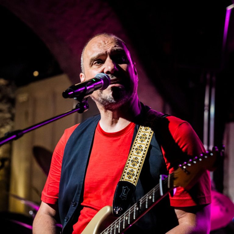 Man met gitaar en microfoon, gekleed in een rode shirt en zwarte vest.
