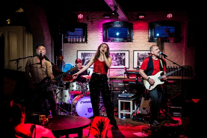 Een band met een vrouwelijke zangeres en muzikanten op het podium tijdens een optreden.