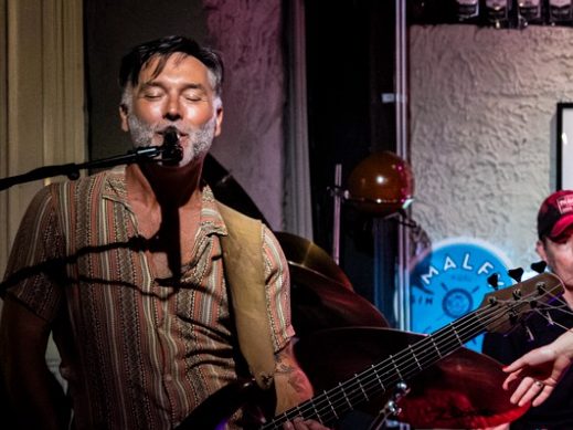 Zanger met gitaar op een podium, omringd door muzikanten en sfeervolle verlichting.