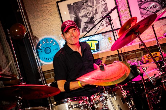 Drummer in een bar speelt op een set percussie met gekleurde verlichting achter zich.