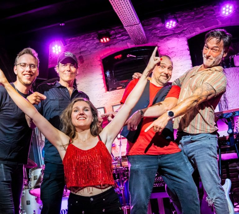Vijf muzikanten poseren enthousiast op een podium met kleurrijke verlichting.