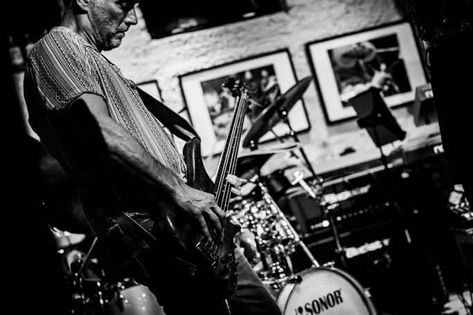 Een muzikant speelt elektrisch gitaar op een podium met een drumstel op de achtergrond.