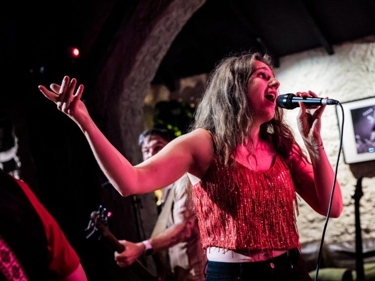 Zangeres optredend met microfoon, terwijl muzikanten op de achtergrond spelen.