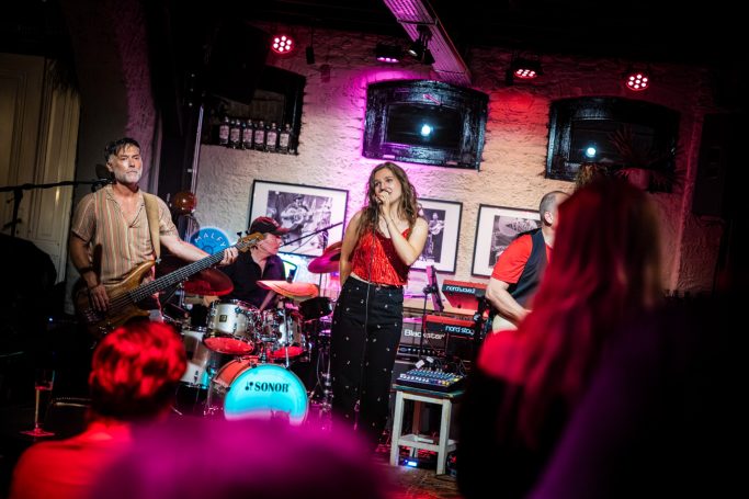 Zangeres optredend met band, met kleurrijke verlichting en publiek op de voorgrond.